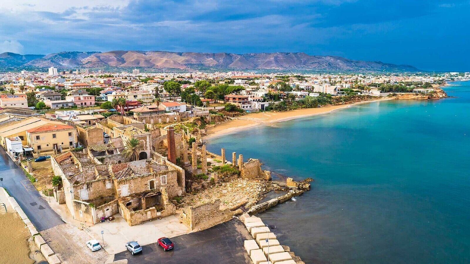 Vue aérienne d’une ville côtière, avec ses bâtiments historiques, sa plage et un paysage montagneux en arrière-plan.