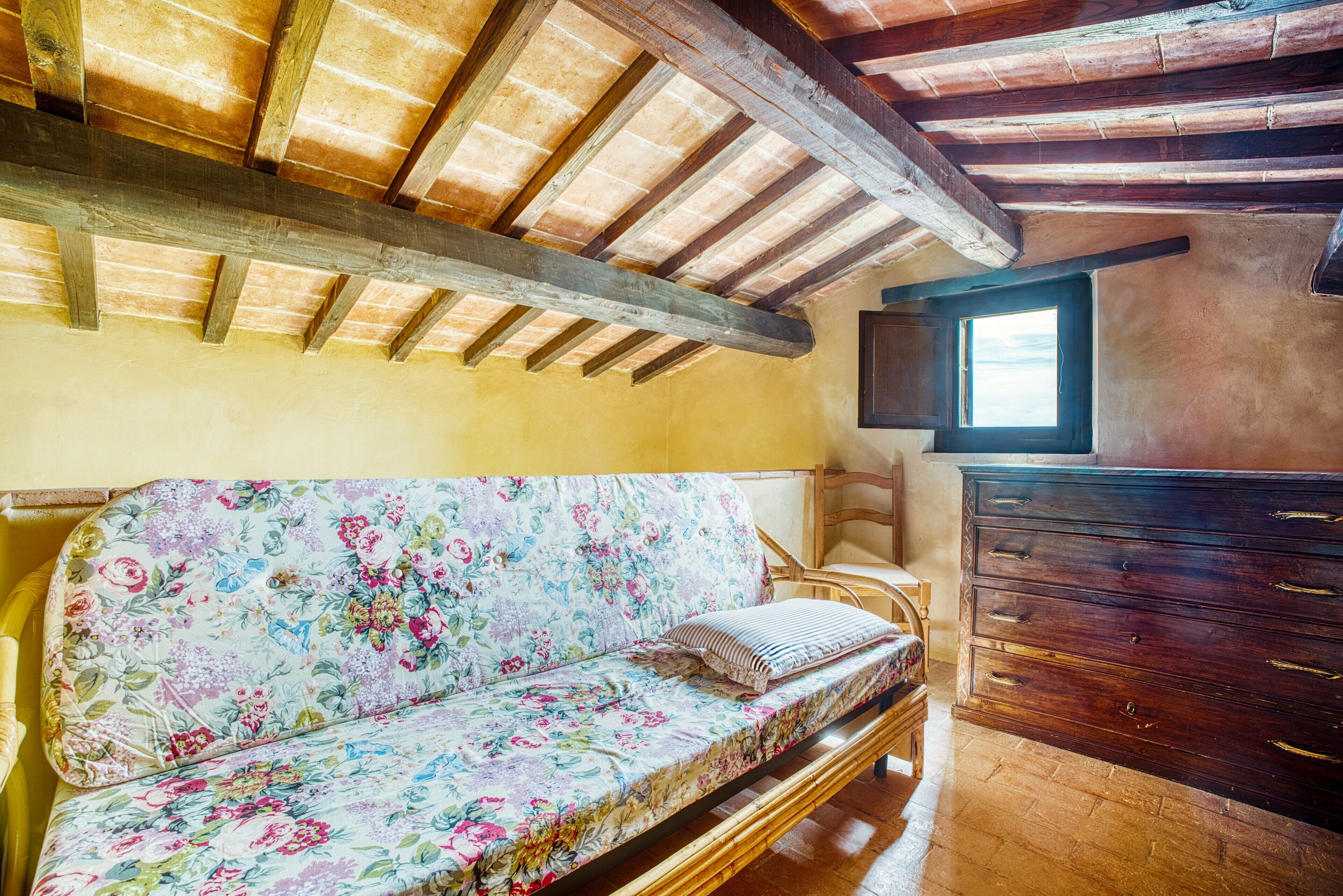 A room with a wooden ceiling, a floral sofa, a wooden dresser, and a window.