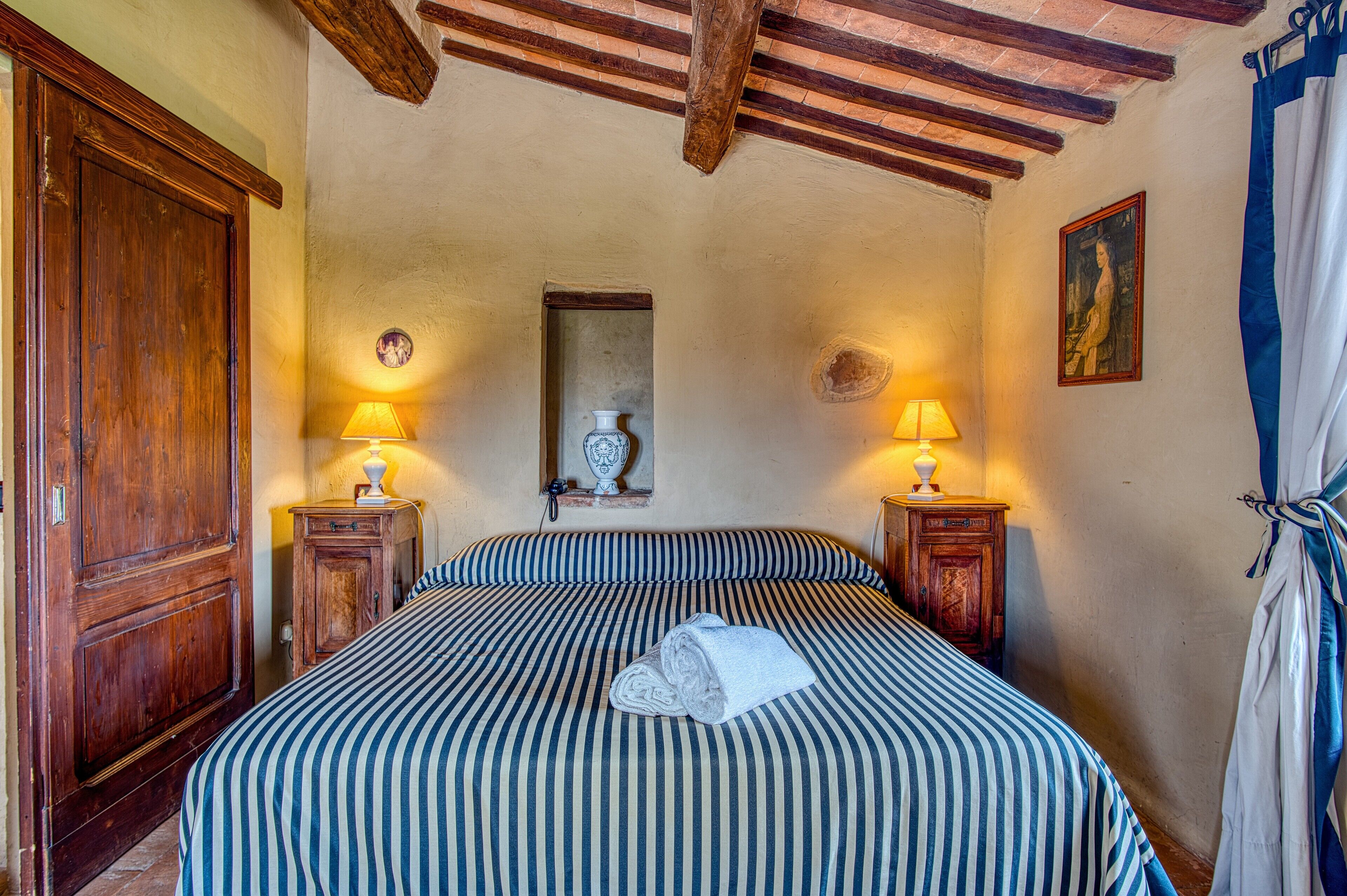A room with a bed, two bedside tables with lamps, a wooden wardrobe, and a painting on the wall.