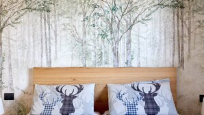 Een slaapkamer met een bed tegen een muur, behangen met een bosmotief, twee kussens met hertenopdruk en een houten hoofdbord.