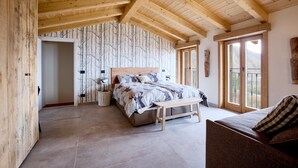 Un dormitorio con techo de madera, cama con ropa de cama estampada, un banco y vista a través de una puerta.