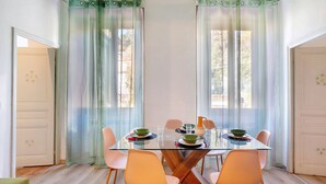 A dining area with a glass table, chairs, and a view through sheer curtains.