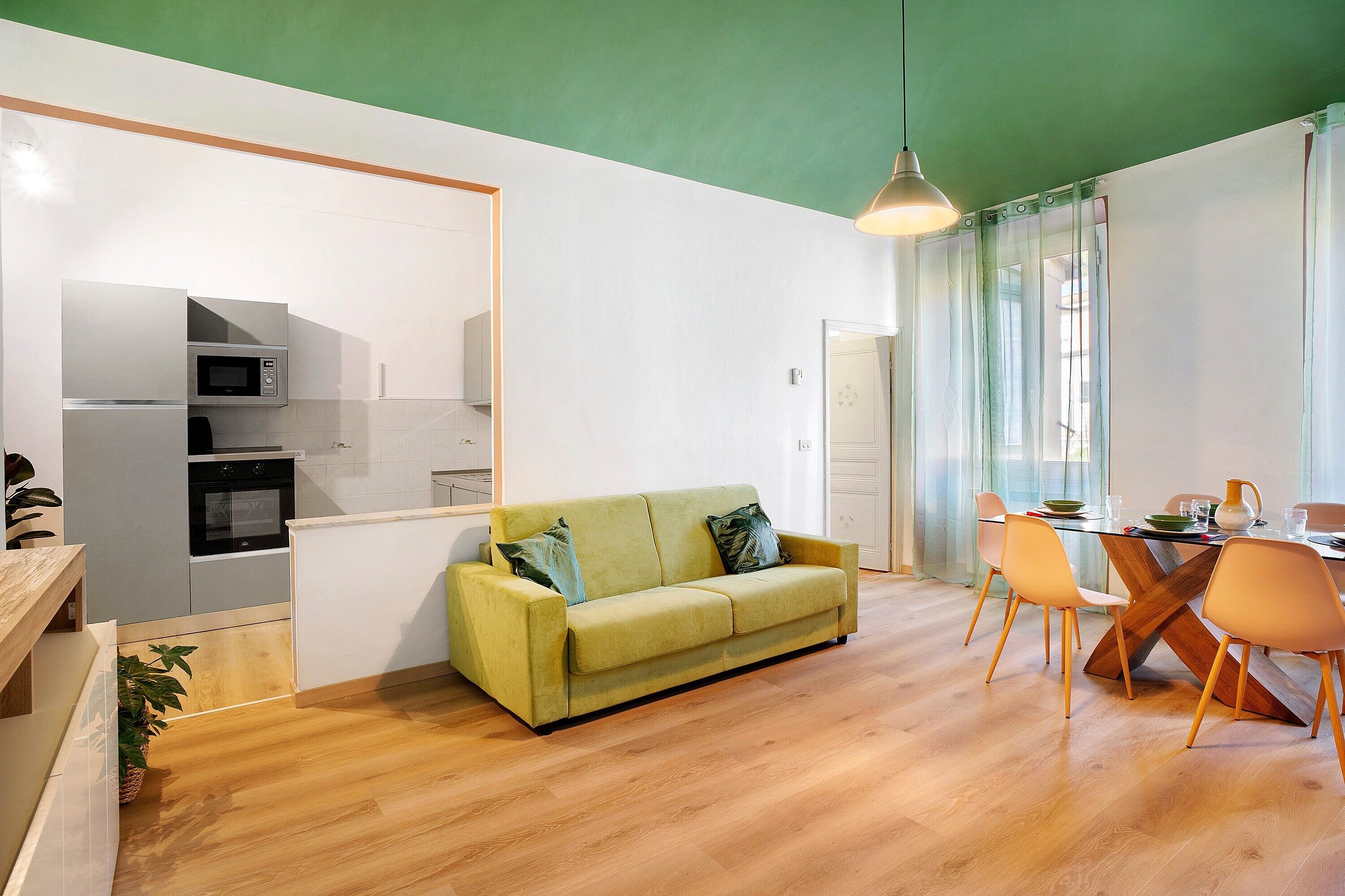 Un espace de vie moderne avec un canapé vert, une table à manger en bois et des chaises, un coin cuisine équipé d’appareils électroménagers et une grande fenêtre avec des rideaux.