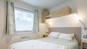 A bedroom with a bed, a window with curtains, a bedside table, and a wall-mounted shelf. - Mobile Home 'Tout Confort Bora Bora' with Shared Pool, Private Terrace and Private Garden (Pont-Aven)