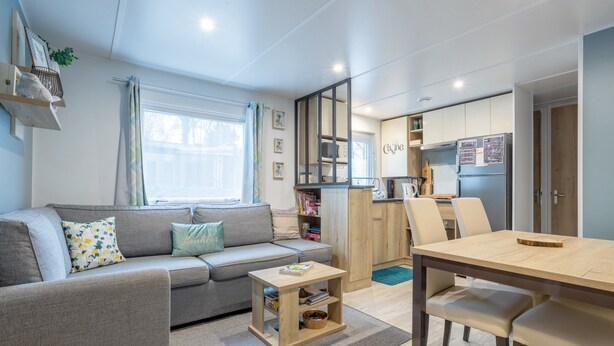 A modern living room with a sofa, a small wooden table, a dining table with chairs, a kitchen area with cabinets and appliances, and a window with curtains. - Mobile Home 'Tout Confort Bora Bora' with Shared Pool, Private Terrace and Private Garden (Pont-Aven)