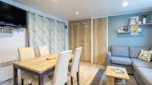 A modern room with a wooden dining table, white chairs, a grey sofa, a TV mounted on the wall, and a wooden wardrobe. - Mobile Home 'Tout Confort Bora Bora' with Shared Pool, Private Terrace and Private Garden (Pont-Aven)