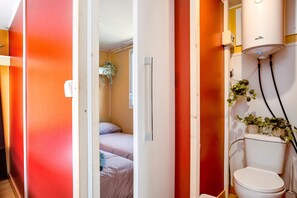 Un bagno con un water, una doccia e un lavandino. Si vede un letto nella stanza adiacente.