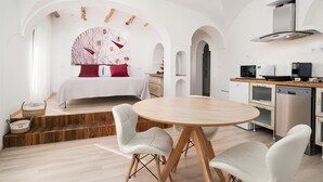 A modern room with a bed, a dining table, white chairs, a kitchen with appliances, and a unique ceiling design.