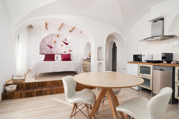 A modern room with a bed, a dining table, white chairs, a kitchen with appliances, and a unique ceiling design.