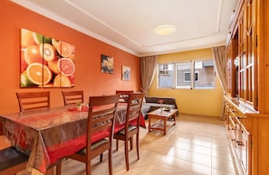 Un espace repas comprenant une table, des chaises et un canapé. On y trouve une fenêtre avec des rideaux, un luminaire au plafond et un mur décoré de peintures représentant des fruits.