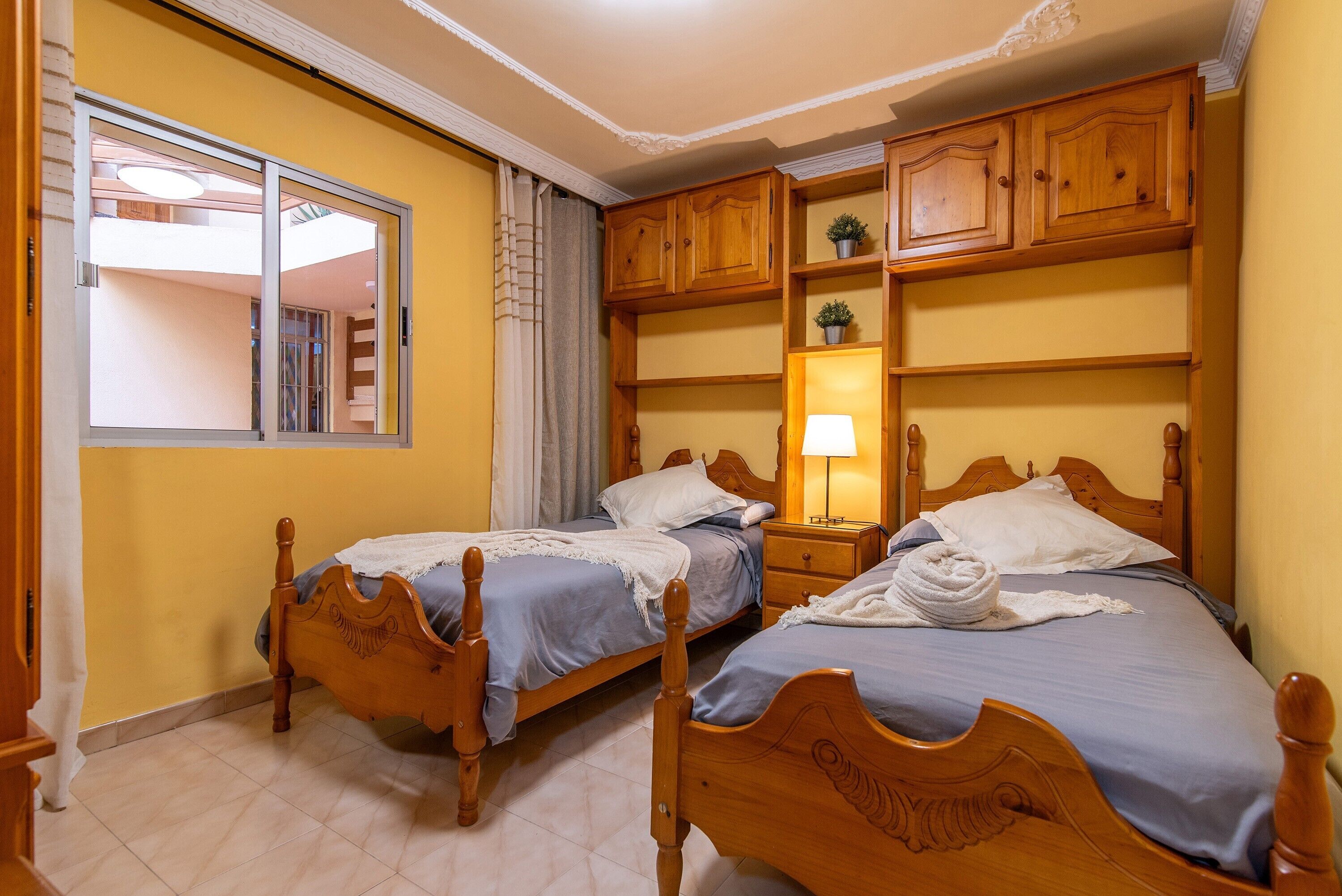 Een slaapkamer met twee houten bedden, een nachtkastje met een lamp, een raam met gordijnen en ingebouwde houten planken.