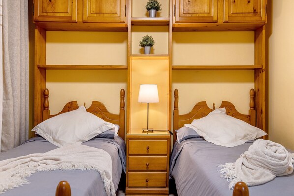 A double room with two beds, wooden headboards, a nightstand with a lamp, and a shelf with a potted plant.