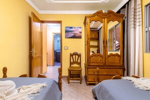 A bedroom with two beds, a wooden wardrobe, a chair, and a painting on the wall.
