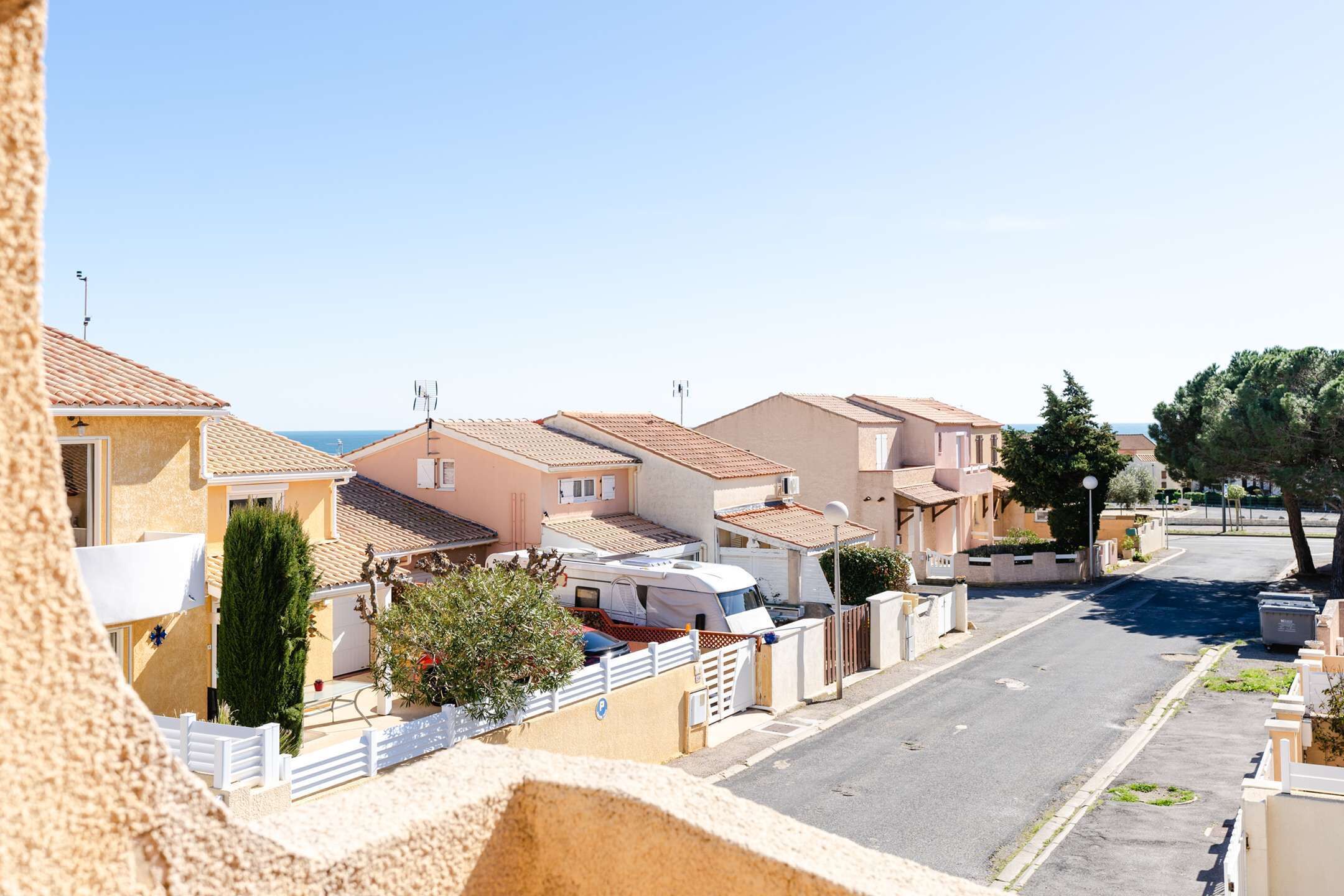 Une rue résidentielle bordée de maisons, avec un camping-car garé et une vue dégagée sur la mer.