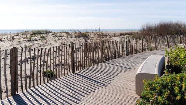 A wooden boardwalk curving along a sandy beach, with a fence on one side and a concrete structure on the other.