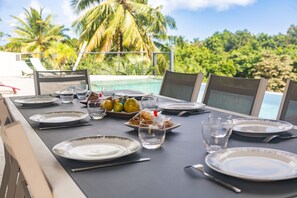 Ein Esstisch für vier Personen, gedeckt mit Tellern, Besteck und Gläsern, mit Blick auf einen Pool und Palmen.