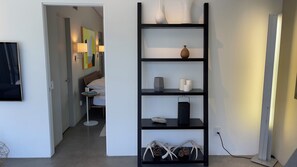 Une chambre d’hôtel moderne avec une étagère noire où sont exposés des objets décoratifs, un lit recouvert de linge de maison blanc et une petite table de chevet avec une lampe.