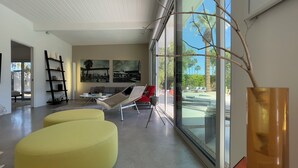 A modern living room with a large glass wall, a yellow ottoman, a red chair, and a black bookshelf.