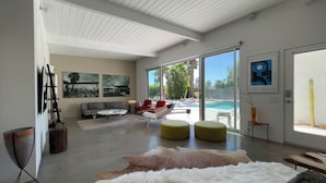 A modern living room with a large glass door leading to an outdoor pool area, a sofa, red chairs, and a yellow ottoman.