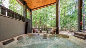 A hot tub with a wooden deck and chairs, surrounded by trees.