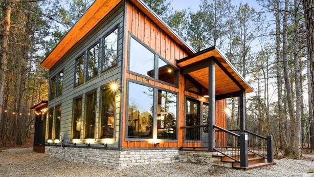 A modern log cabin with large windows, a wooden deck, and a stone foundation.