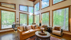 A spacious living room with a large sofa, a round coffee table, and a fireplace. There are multiple windows with blinds, and a view of trees outside.