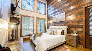 A bedroom with a large bed, wooden walls, and a view of trees through the windows.