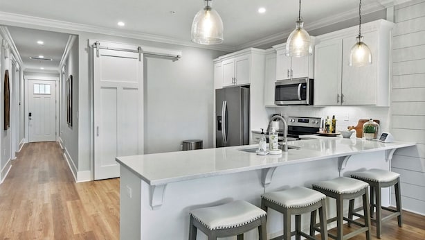 A modern kitchen with white cabinetry, a large island with bar stools, stainless steel appliances, and pendant lighting. - Moorings 104 Beautiful Marsh Front Condo (Jekyll Island)