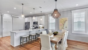 A modern kitchen with white cabinetry, a central island, and a dining area with a wooden table and bar stools. - Moorings 104 Beautiful Marsh Front Condo (Jekyll Island)