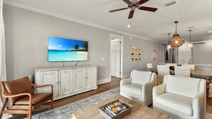 A spacious living room with a ceiling fan, a flat-screen TV displaying a beach scene, a dining area with a wooden table, and a kitchen in the background. - Moorings 104 Beautiful Marsh Front Condo (Jekyll Island)