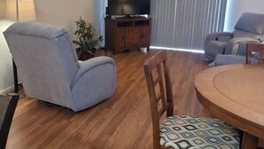 A living room with a wooden dining table, a grey armchair, a patterned chair, a television on a wooden stand, and a ceiling fan. - 1st Floor Condo in the Desert w/ Pool, Spa & BBQ (Lake Havasu City)