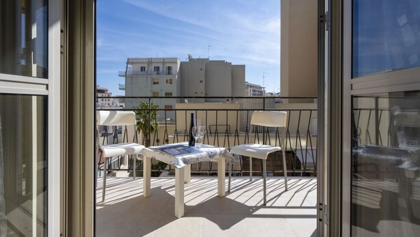 A balcony with a table and chairs, a glass door, and a view of buildings. - GuestHost - Large and bright apartment that can accommodate up to 5 people with 2 bedrooms and 2 bathrooms, which boasts a beautiful view of the Villa di Sant'Antonio and, in the distance, of the sea from the balcony and window. Located a few steps from  (Monopoli)