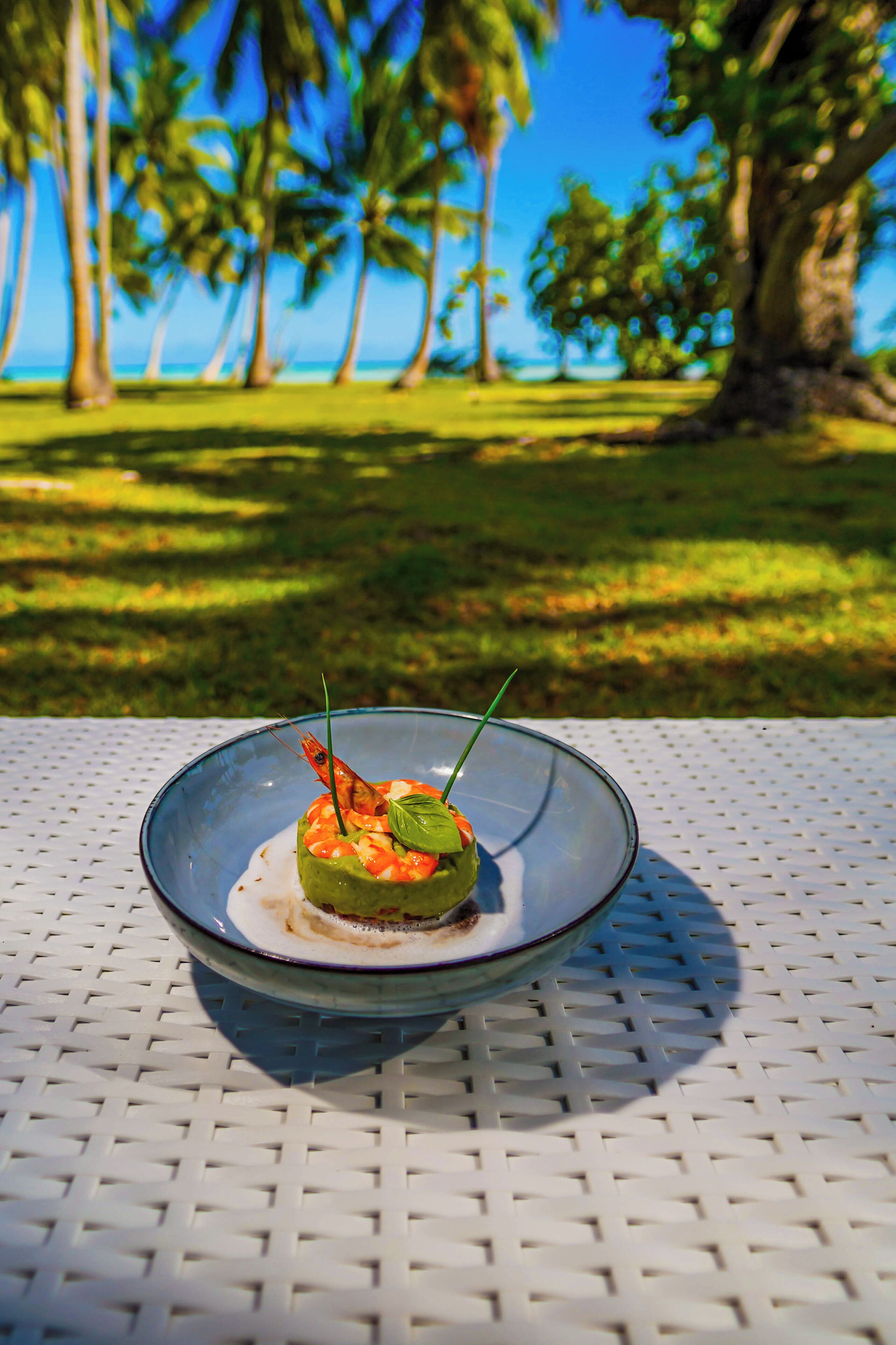 A clear view of a dining table with a glass bowl containing a dish, set against a backdrop of palm trees and a clear sky.