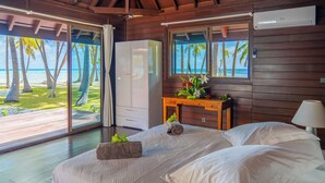 Une chambre avec une grande fenêtre, des murs en bois, un lit avec du linge de lit blanc, une table de chevet en bois et une vue sur la plage.