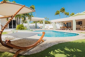 A pool area with a wooden lounge chair, a covered seating area, and a view of palm trees. - White Lotus Retreats (Noord)