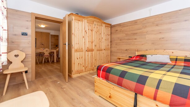 A bedroom with a wooden wardrobe, a bed with a colorful checkered bedspread, a wooden chair, and a wooden desk with a chair. - Casa Caviola Centro (Falcade)