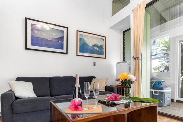 Un salon avec un canapé gris foncé, deux tableaux représentant des paysages encadrés, une table en verre avec une bouteille, deux verres et un vase de fleurs.