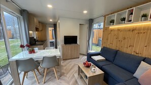 A modern living room with a sofa, a dining table with chairs, a TV, and a kitchen area with wooden cabinets and a window. - Villa Ramses by Interhome (Riedenburg)