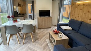 A modern, well-lit living room with a sofa, a dining table with chairs, a TV, and a kitchen area with cabinets and a window. - Villa Ramses by Interhome (Riedenburg)