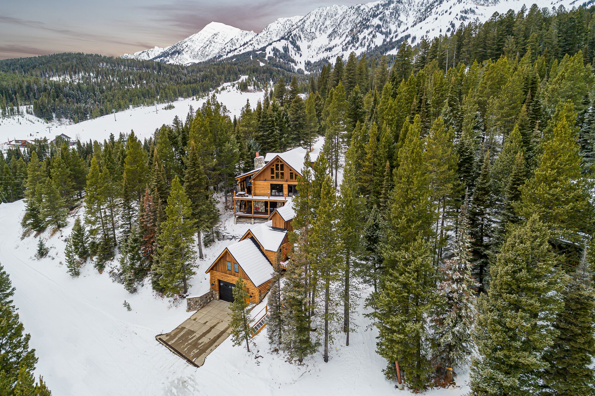 Una cabaña de madera con un porche grande y un garaje, rodeada de nieve y un bosque denso, con montañas al fondo.