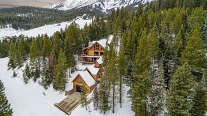 A wooden cabin with a large porch and a garage, surrounded by snow-covered ground and dense forest, with mountains in the background. - Bridger Lodge: Ski-In Ski-Out/Hike/Bike/Fish/Hunt Bridger Bowl Ski Area (Bozeman)