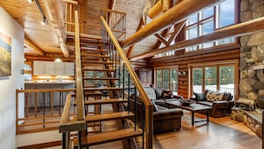 A spacious log cabin interior with a wooden staircase, a stone fireplace, and large windows offering views of the outdoors. - Bridger Lodge: Ski-In Ski-Out/Hike/Bike/Fish/Hunt Bridger Bowl Ski Area (Bozeman)
