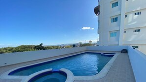 A rooftop swimming pool with a hot tub, surrounded by a tiled deck, and a view of a cityscape.