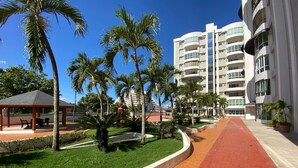 A hotel corridor with palm trees, a covered seating area, and a red pathway.