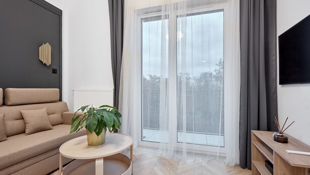 A modern living room with a sofa, a round table with a plant, a TV on a wooden stand, and a large window with curtains. - Nadolnik Apartment with Mezzanine (Poznan)