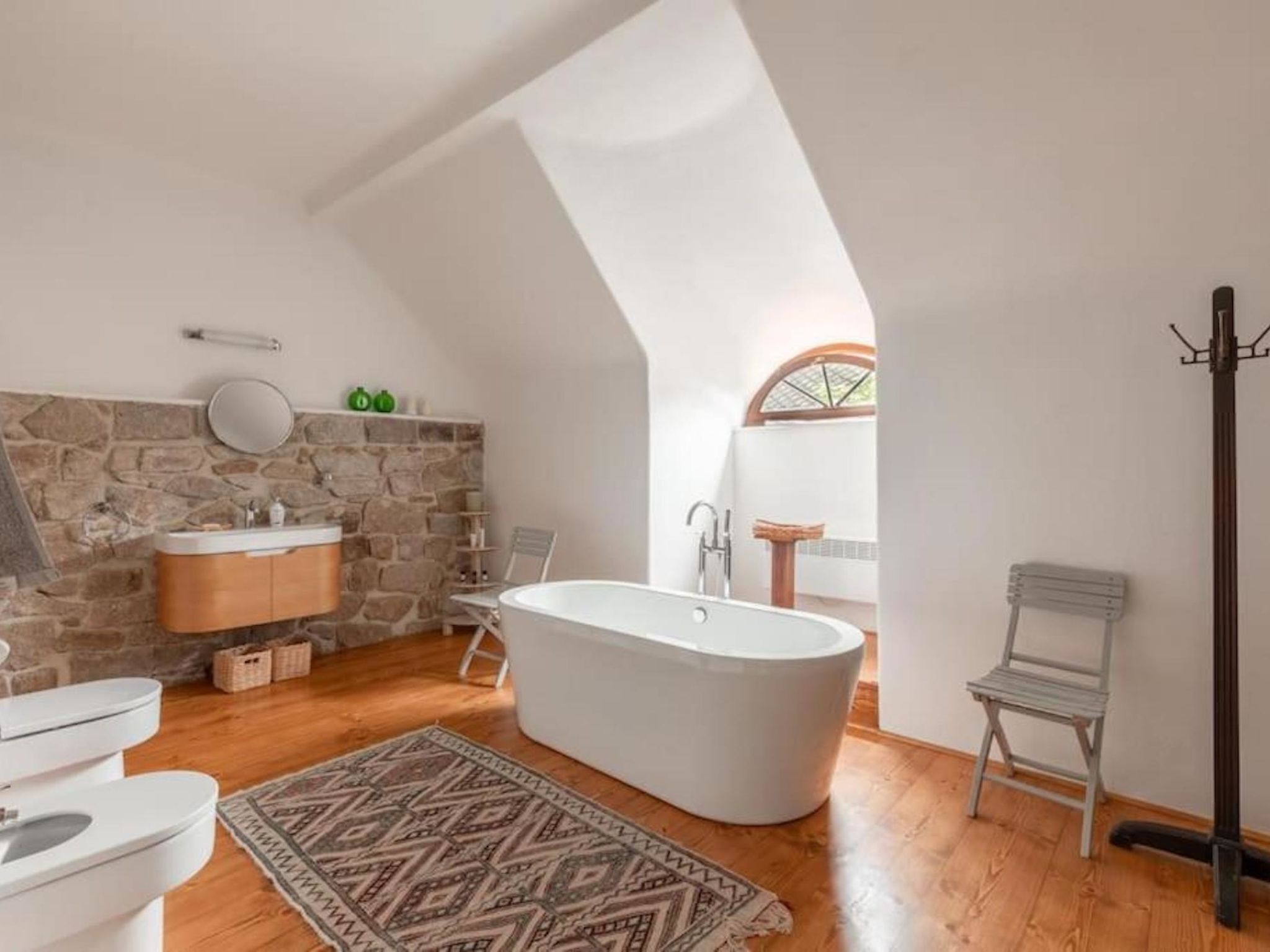 A bathroom with a stone wall, a freestanding bathtub, a white sink, and a wooden floor.