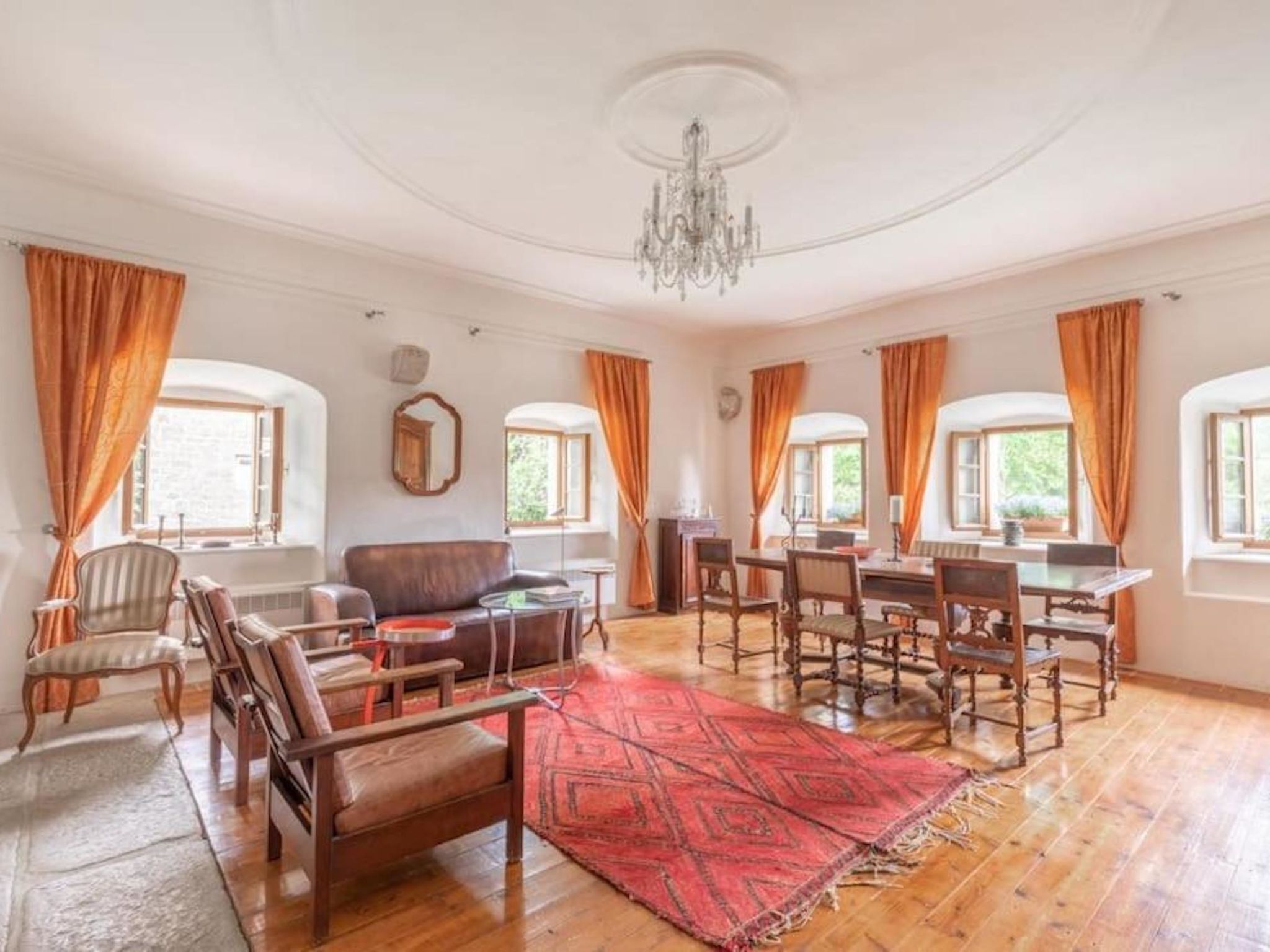 A spacious living room with a large wooden dining table, several chairs, a sofa, and a red patterned rug.