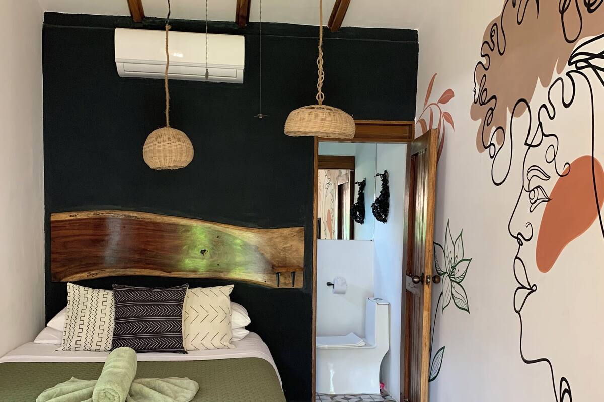 A bedroom with a wooden bed, a green bedspread, a headboard, two hanging lights, and a wall mural.