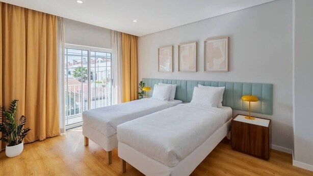 Habitación de hotel con dos camas, un ventanal grande con cortinas, una planta en maceta y una mesita de noche con lámpara.