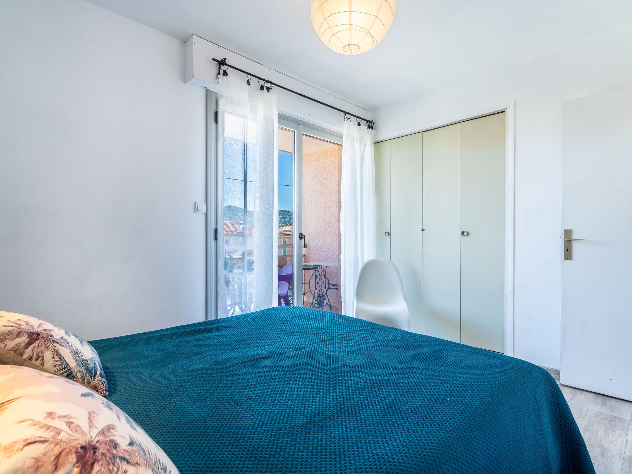 A bedroom with a bed, a chair, a wardrobe, and a sliding glass door leading to a balcony.